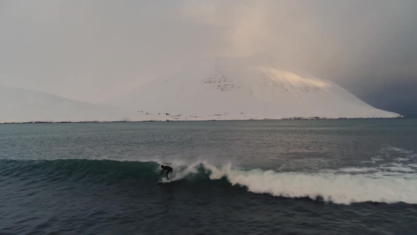 Aerial view of a person surfing a wave in a winter ocean with snowy mountains on the horizon