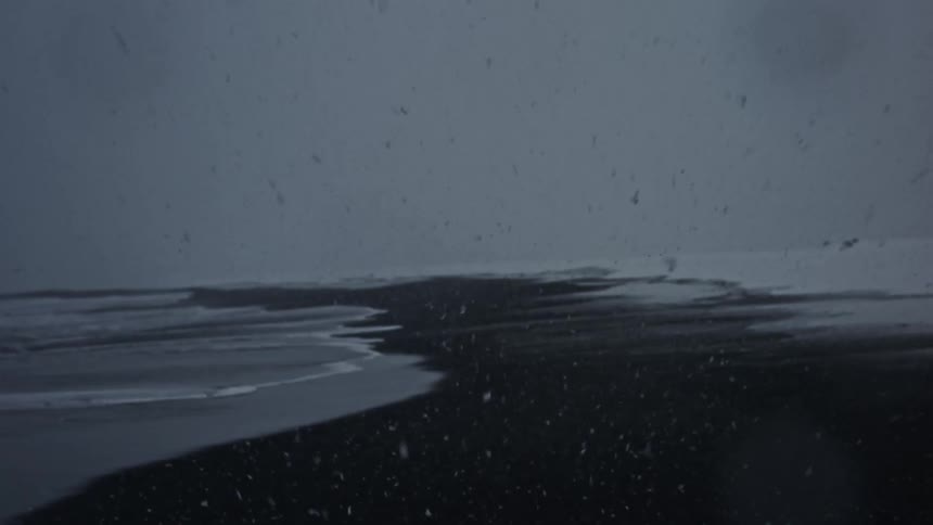 A dark snowy beach with waves gently lapping against the shore and falling snow under a gray overcast sky