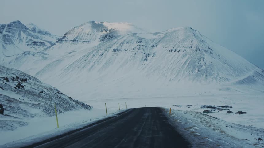 A snowcovered mountain range with a road leading towards it flanked by yellow markers on both sides