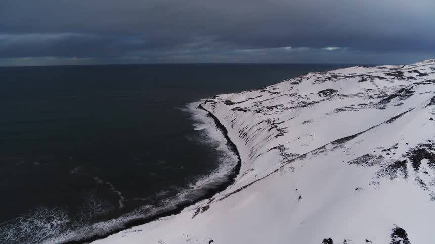A snowcovered hillside slopes down to a dark rugged coastline bordering a turbulent sea under a dramatic sky