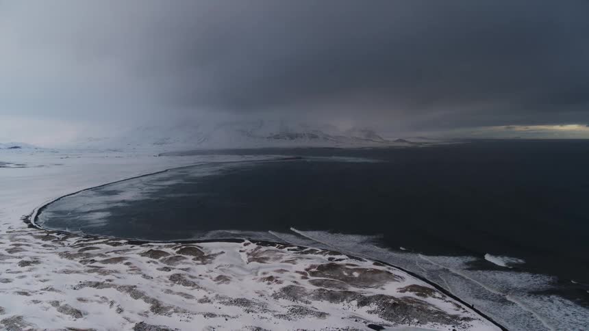 A dark curving shoreline meets a vast snowcovered landscape under an ominous stormy sky