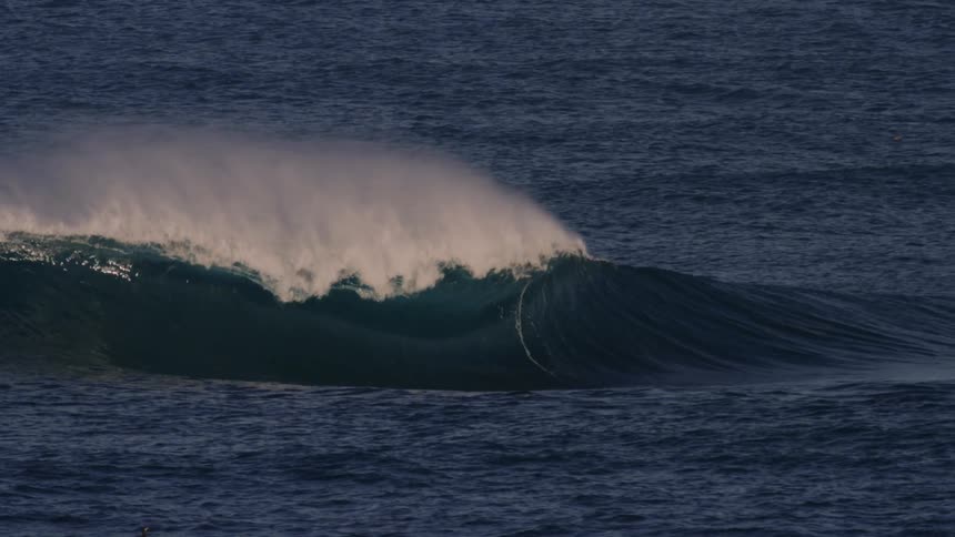 A large wave curls and breaks in the deep blue ocean forming a whitecap against the backdrop of the vast sea