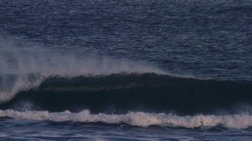 A large ocean wave is cresting and breaking with mist spraying off the top against a backdrop of deep blue water