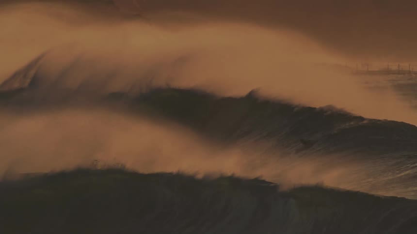 A massive wave crashes in a swirl of foam and spray illuminated by a warm golden light with surfers dwarfed by the size of the wave