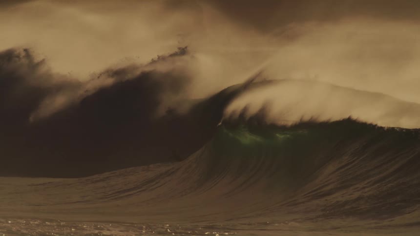 A massive wave crashes in a swirl of foam and spray illuminated by a warm golden light against a hazy sky with surfers dwarfed by the size of the wave