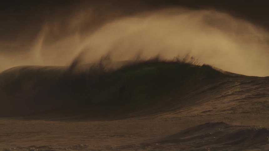 A massive wave crashes in a swirl of foam and spray illuminated by a warm golden light with surfers dwarfed by the size of the wave