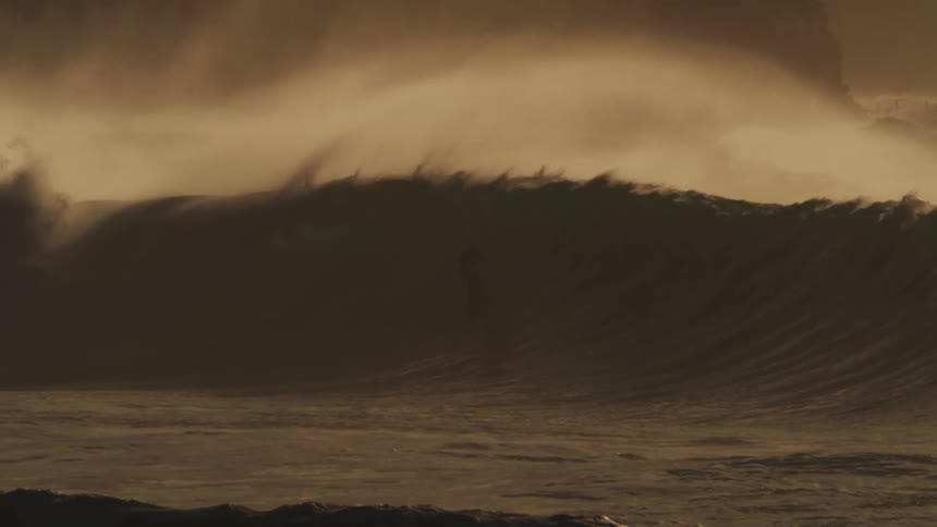 A massive wave crashes in a swirl of foam and spray illuminated by a warm golden light against a hazy sky with surfers dwarfed by the size of the wave