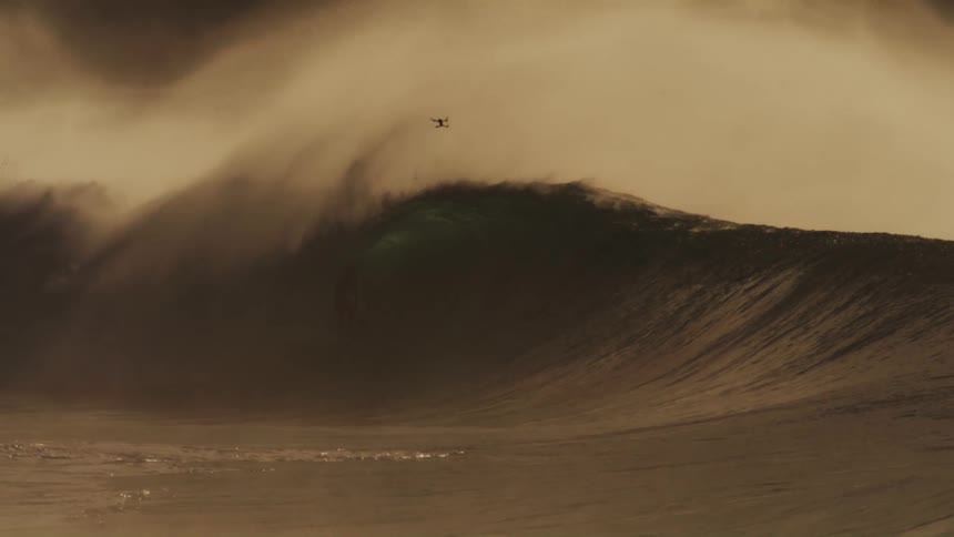 A massive wave crashes in a swirl of foam and spray illuminated by a warm golden light against a hazy sky with surfers dwarfed by the size of the wave