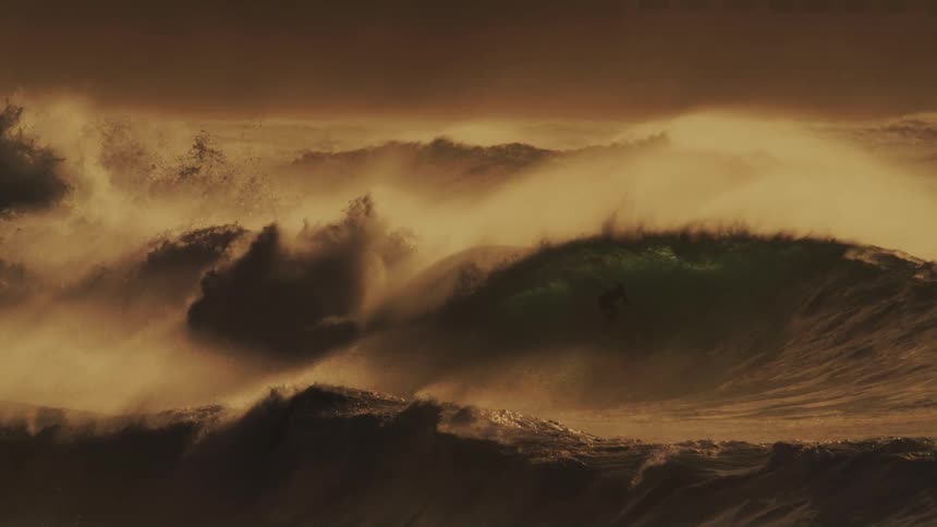 A massive wave crashes in a swirl of foam and spray illuminated by a warm golden light with surfers dwarfed by the size of the wave