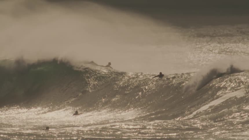 A massive wave crashes in a swirl of foam and spray illuminated by a warm golden light against a hazy sky with surfers dwarfed by the size of the wave