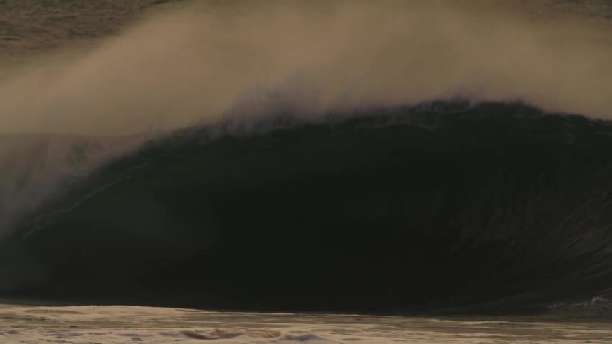 A massive wave crashes in a swirl of foam and spray illuminated by a warm golden light against a hazy sky with surfers dwarfed by the size of the wave