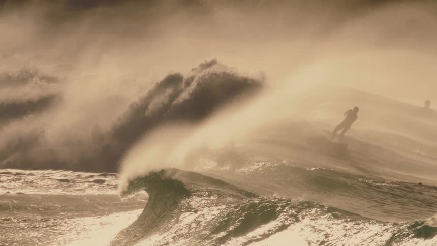 A powerful wave breaks in a sepia toned seascape with surfers visible in the background