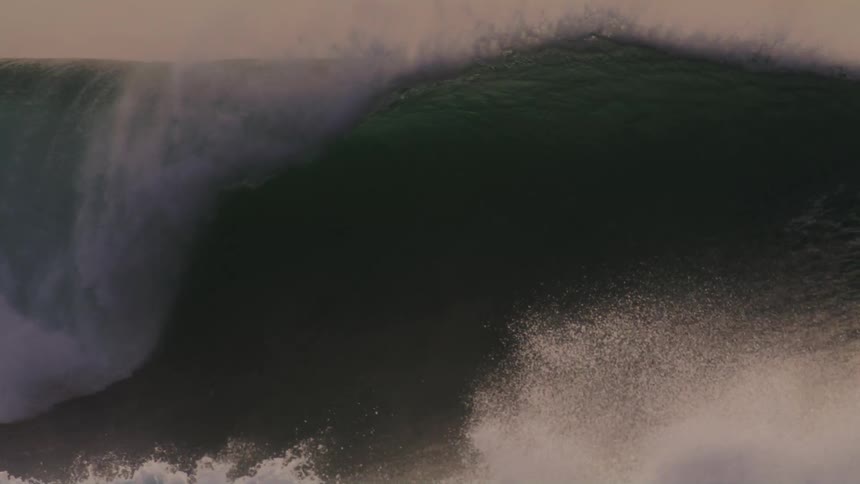 Surfers on top of a towering wave while another surfer watches from the water