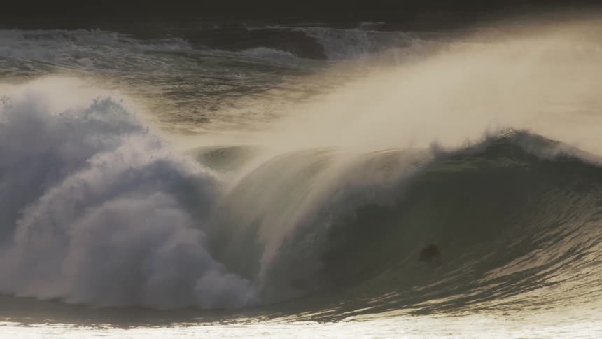 A massive wave crashes in a swirl of foam and spray illuminated by a warm golden light against a hazy sky with surfers dwarfed by the size of the wave