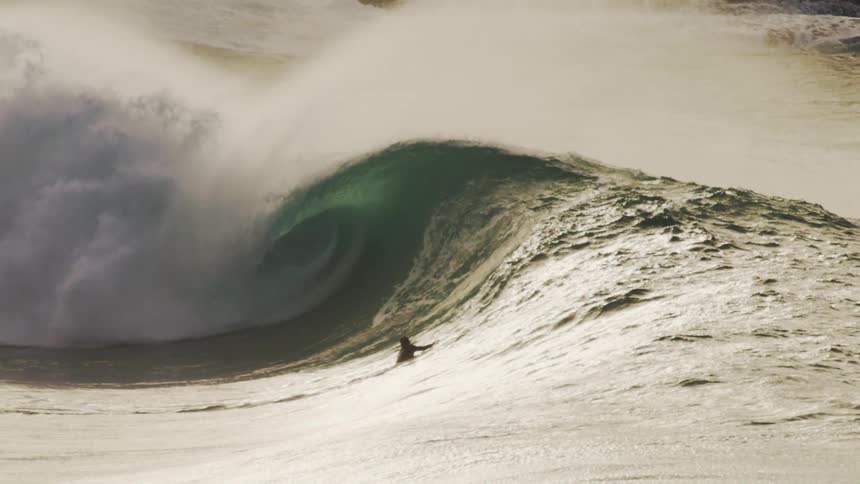 A massive wave crashes in a swirl of foam and spray with surfers dwarfed by the size of the wave