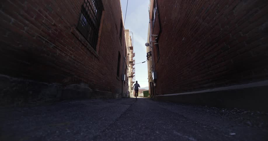 A person is running down a narrow alleyway between two brick buildings with the perspective looking up from the ground towards the sky