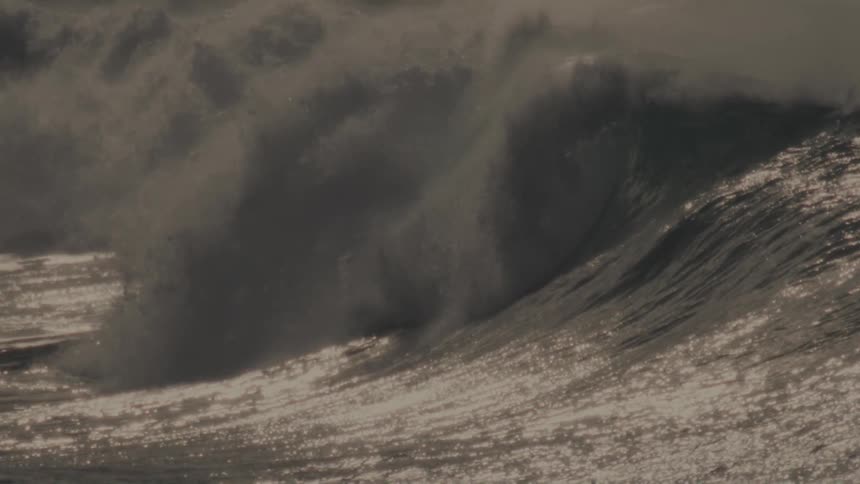 A massive wave crashes in a swirl of foam and spray illuminated by a warm golden light with surfers dwarfed by the size of the wave