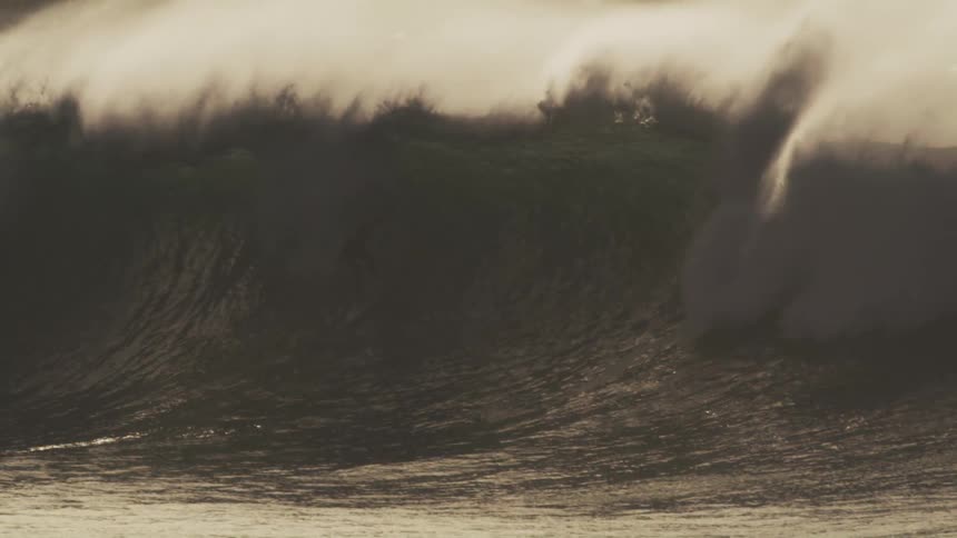 A massive wave crashes in a swirl of foam and spray illuminated by a warm golden light against a hazy sky with surfers dwarfed by the size of the wave