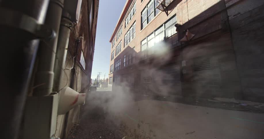 Jogger runs through an alley between warehouses during the day passing a port emitting vapor