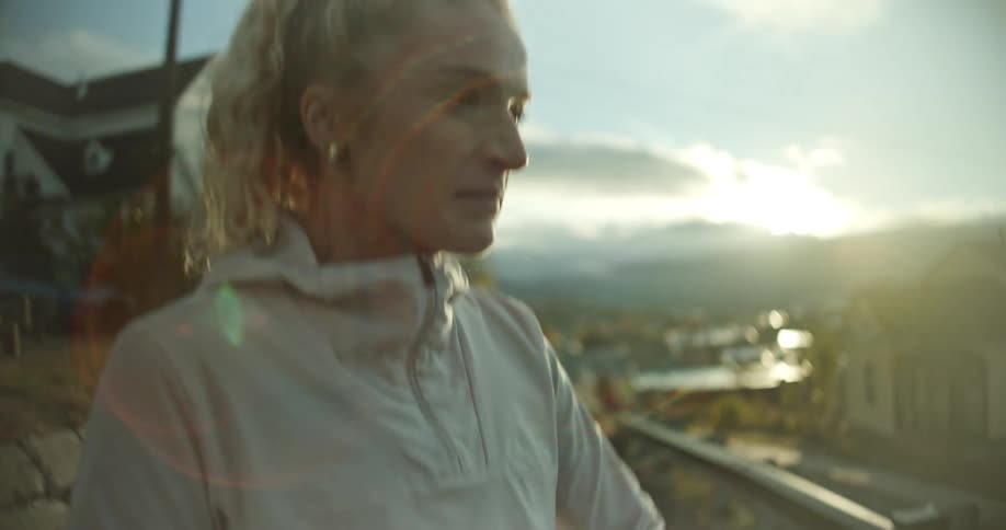 A woman in a light jacket checks her wristwatch on a road overlooking a scenic town with mountains at sunrise or sunset