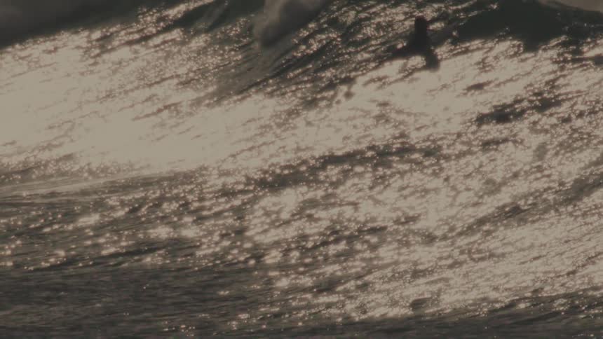 A massive wave crashes in a swirl of foam and spray with surfers dwarfed by the size of the wave