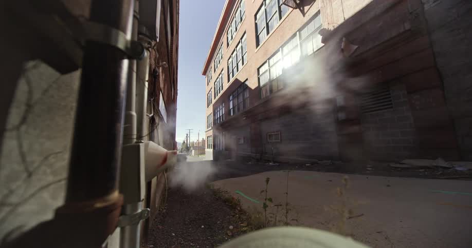 A woman in a grey hoodie and shorts runs through steam venting from an exterior wall in an alley
