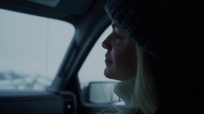 A woman wearing a winter hat looks out the window of a car driving through a snowy landscape