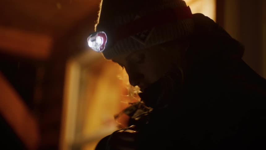 A woman wearing a headlamp illuminated in a dimly lit environment adjusts a thick winter jacket