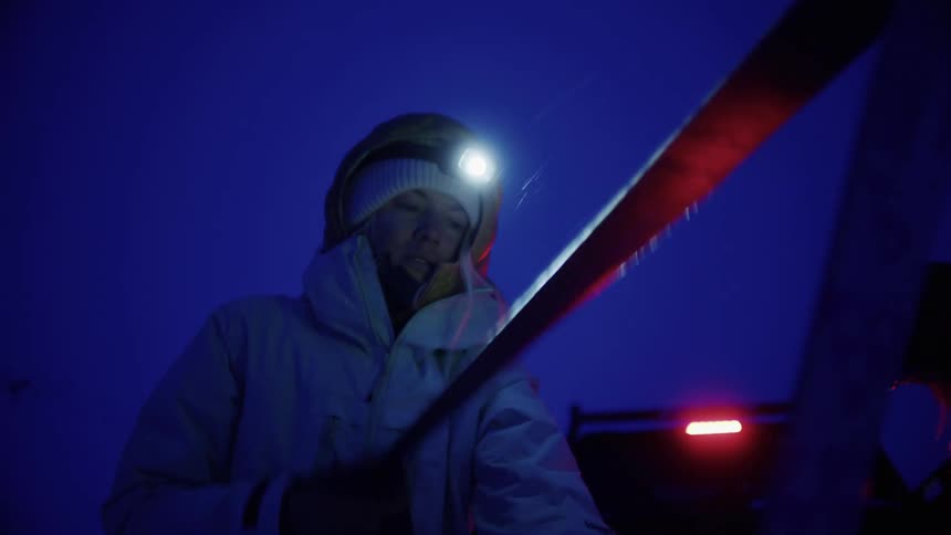 Woman preps her ski at night while standing with another person near a vehicle parked in the snow