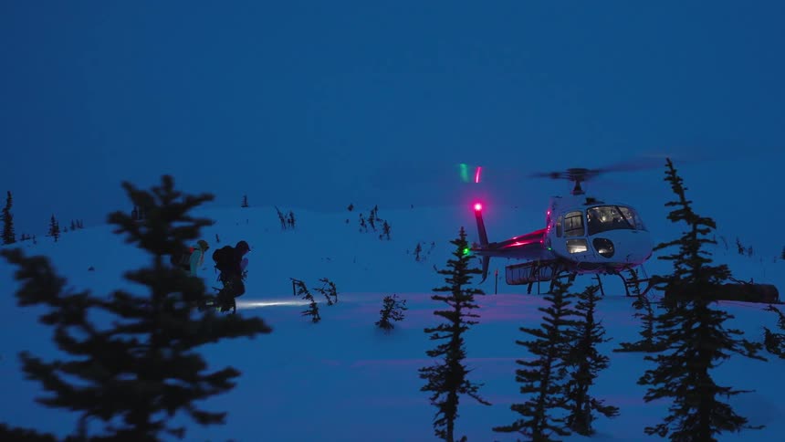 Two skiers approach a helicopter on a snowy mountain at dusk illuminated by the helicopters lights