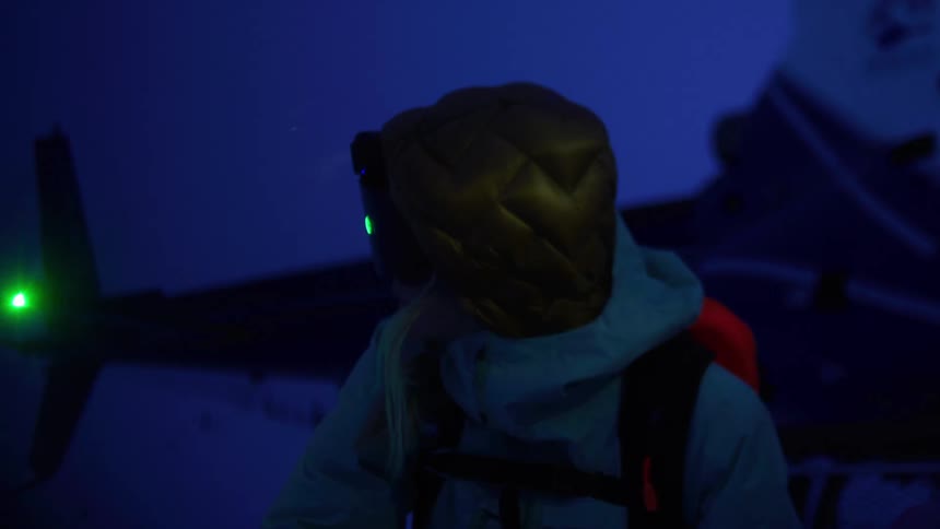 A person wearing a headlamp and winter clothing stands in a dark snowy environment with a helicopters spinning rotor in the background