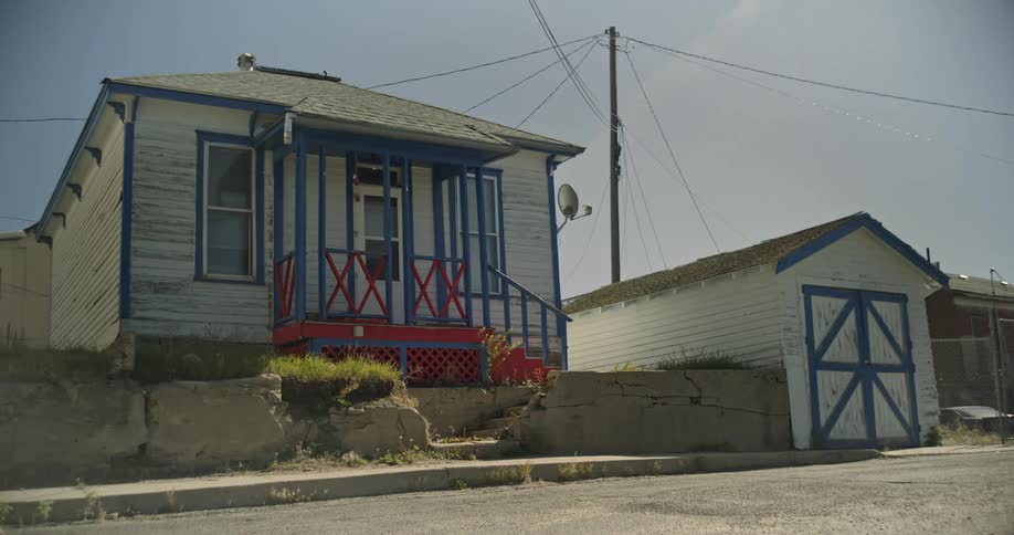 A woman runs along a sidewalk past a small weathered house with blue trim and a detached garage under a bright sky