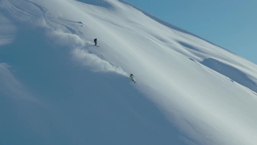 Two skiers are descending a steep snowcovered mountain slope leaving trails of powdery snow behind them under a clear blue sky