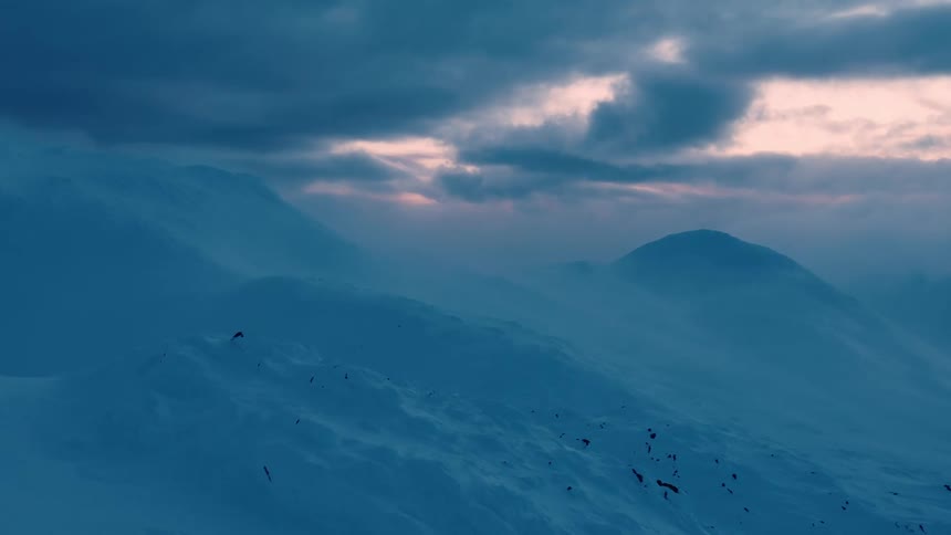 Fog hovers over snowy mountainous terrain and under the dark clouds in a red tinted sky
