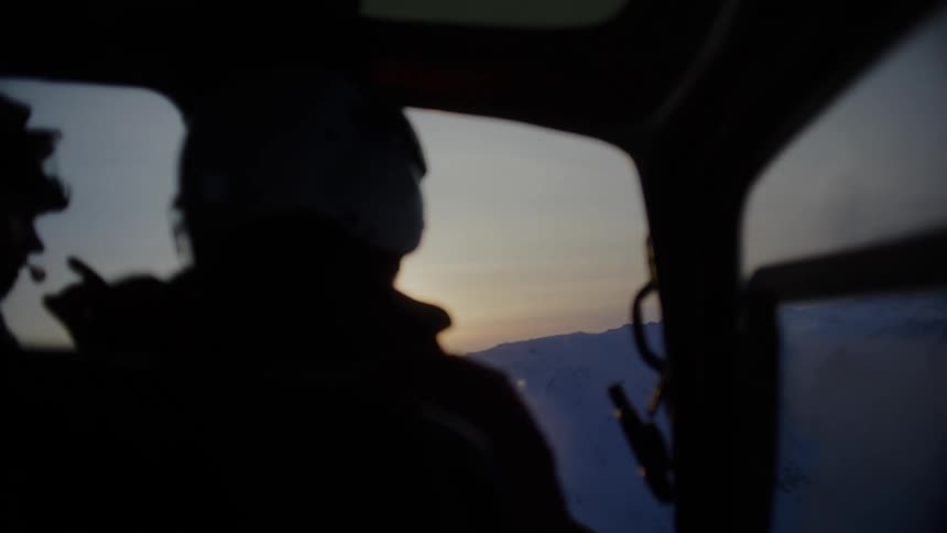 The silhouette of a pilot is seen in the cockpit of a helicopter against a backdrop of snow covered mountains and a sunset