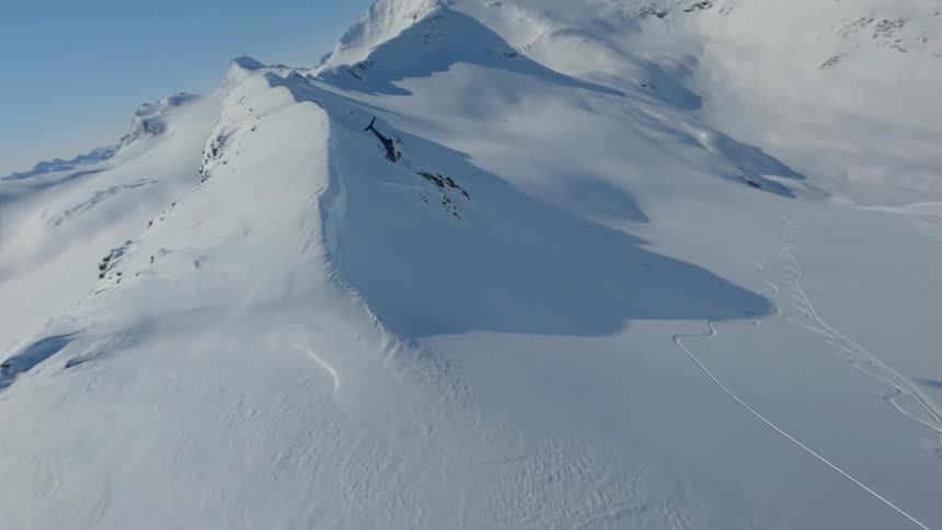A helicopter flies over a snowcovered mountain range near a snowboarder wearing a red jacket