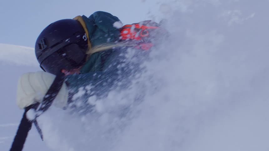 A skier in a green jacket and light blue pants is skiing through deep snow creating a spray of snow behind while holding ski poles and wearing a helmet and goggles