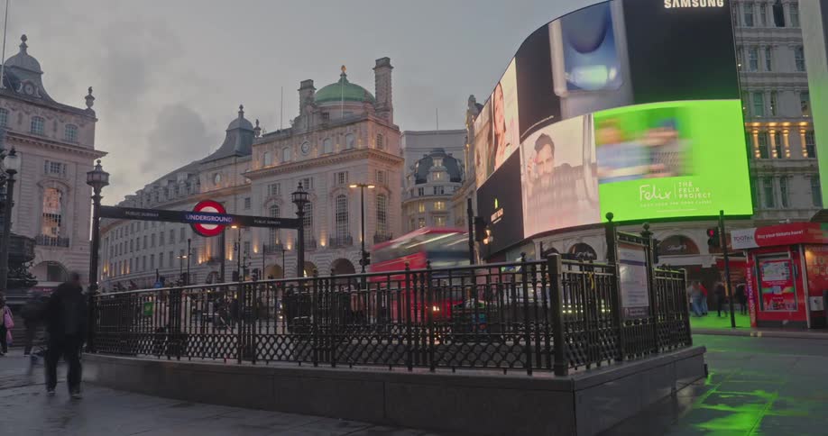 A red doubledecker bus passes pedestrians by the iconic curved advertising screens and historic buildings of piccadilly circus in london under an overcast sky