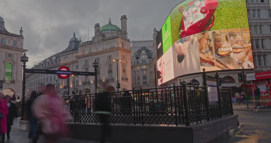 Red doubledecker buses and pedestrians pass by the iconic curved advertising screens and historic buildings of piccadilly circus in london under an overcast sky