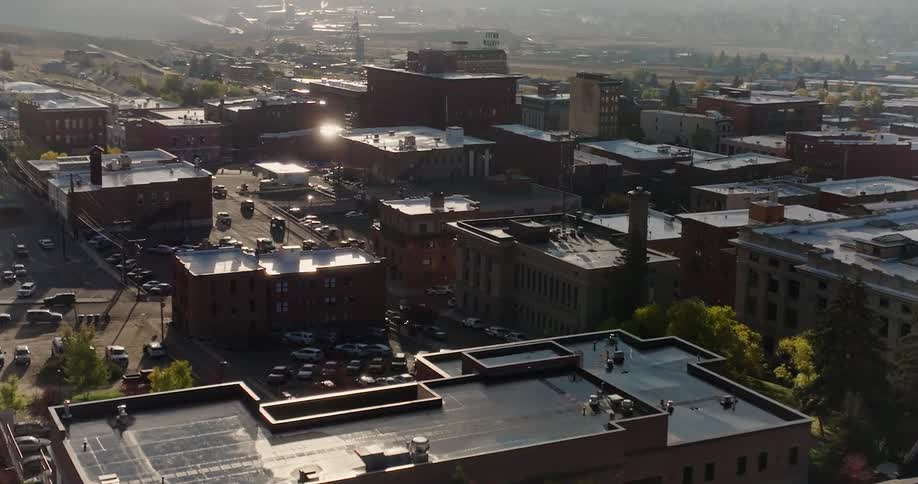 An aerial view of a cityscape with numerous buildings streets and parked cars under a hazy sky with sunlight reflecting off some rooftops