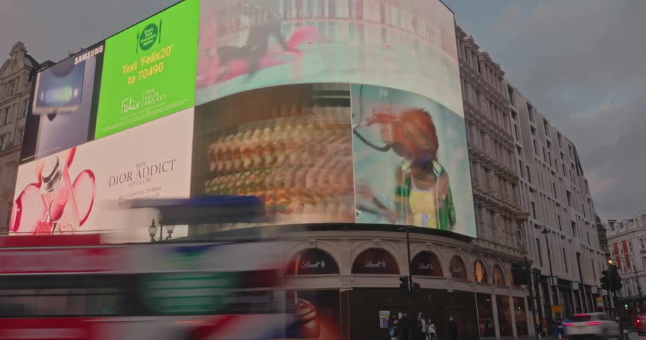 Red doubledecker buses and pedestrians pass by the iconic curved advertising screens and historic buildings of piccadilly circus in london under an overcast sky