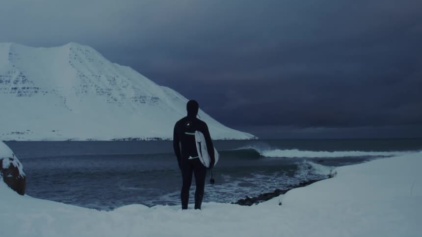 Person in swimsuit holding a surfboard on a snowy mountain facing the sea