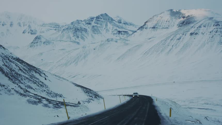 A white suv drives down a paved road winding through a vast snowcovered mountain landscape under a cloudy sky