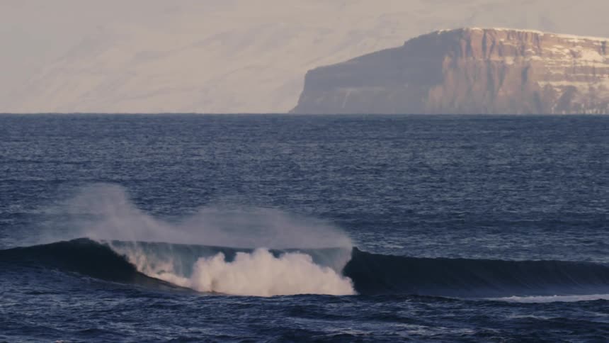 A wave curls and breaks in the ocean under a hazy sky with a large flattopped mesa visible in the distance