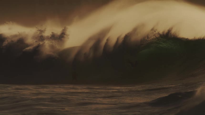 A massive wave crashes in a swirl of foam and spray illuminated by a warm golden light against a hazy sky with surfers dwarfed by the size of the wave