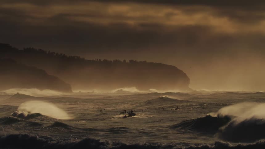 A jetski transports surfers through an ocean with massive crashing waves foam and spray