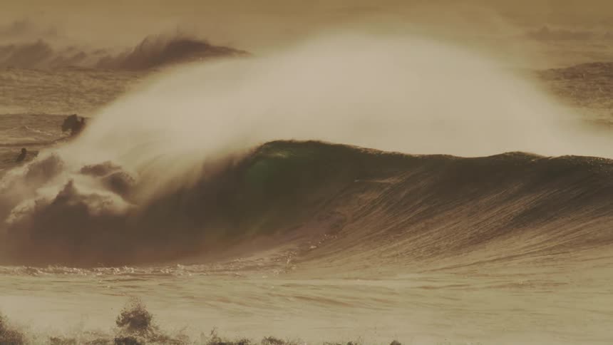 A massive wave crashes in a swirl of foam and spray illuminated by a warm golden light against a hazy sky with surfers dwarfed by the size of the wave