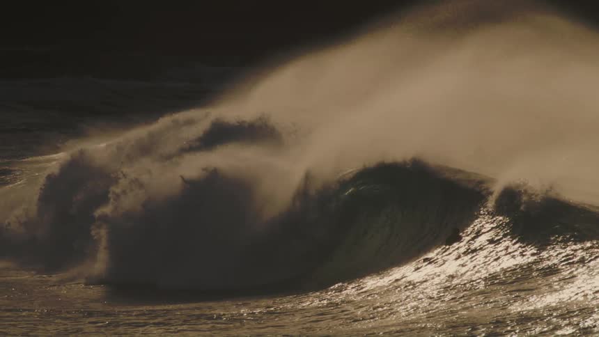 A massive wave crashes in a swirl of foam and spray illuminated by a warm golden light with surfers dwarfed by the size of the wave