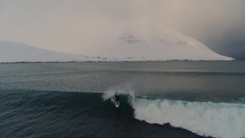 A lone surfer rides a wave in a cold expansive ocean with a snowcovered mountain in the background