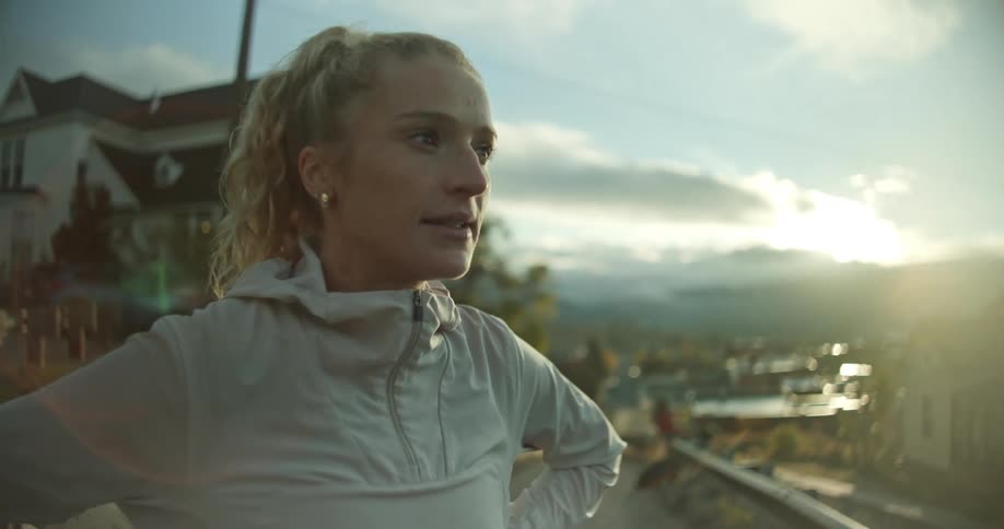 A woman in a light jacket stands on a road overlooking a scenic town view with houses trees and mountains under a setting or rising sun